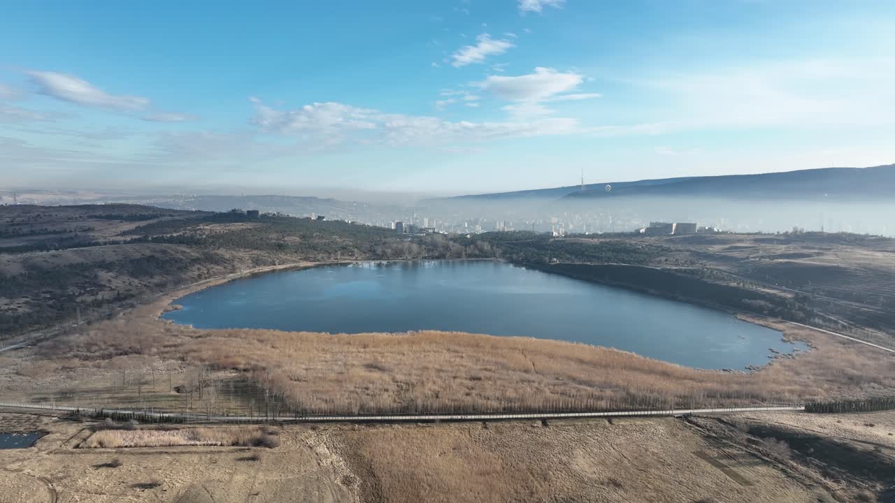 A breathtaking aerial view of a tranquil lake with a vast open field surrounding it. The reeds add a golden frame to the deep blue waters, and a few distant structures can be spotted near the horizon