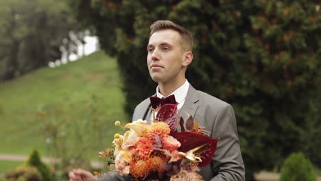 Groom-goes-down-the-alley-between-bushes-with-a-wedding-bouquet-to-his-beloved-bride,-first-meeting