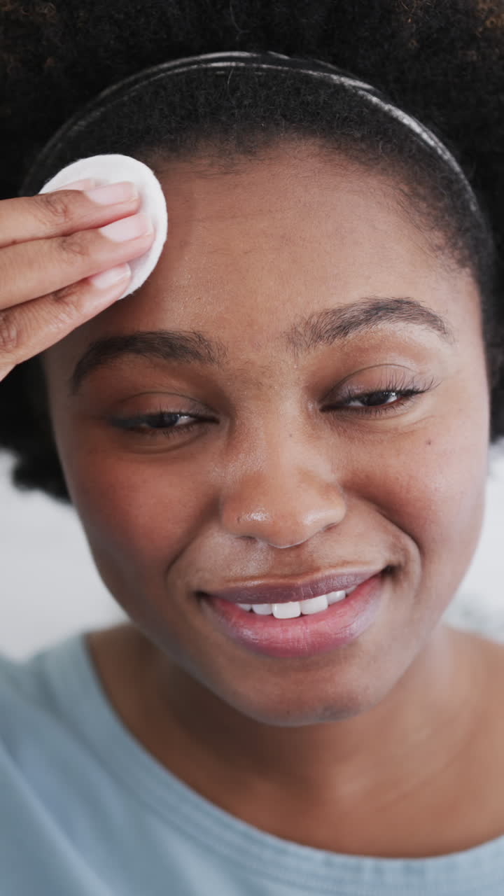 Vertical video of portrait of african american woman cleaning face with cotton pad
