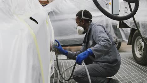 African-American-male-car-mechanic-wearing-a-face-mask-and-using-a-grinder-on-the-side-of-a-car