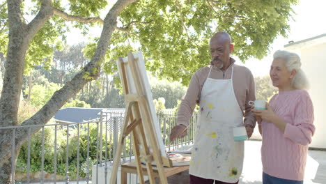 happy senior biracial couple painting and drinking coffee at balcony at home, slow motion