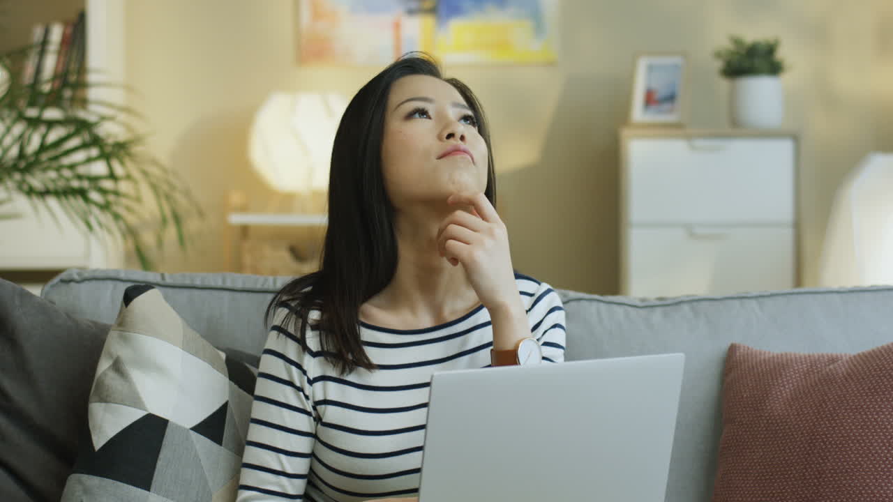 Young Woman In Striped Shirt Working With Laptop On Laps And Thinking Sitting On The Sofa In The Living Room
