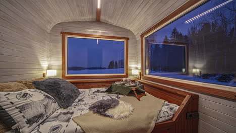 vista estática de un acogedor dormitorio en una cabaña de madera con una enorme ventana de vidrio que da al campo cubierto de nieve al amanecer en un lapso de tiempo