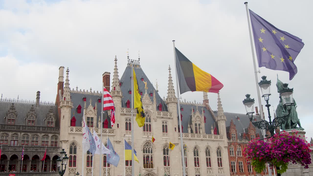 Multiple national and regional flags wave in wind before ornate medieval building, daylight, static shot