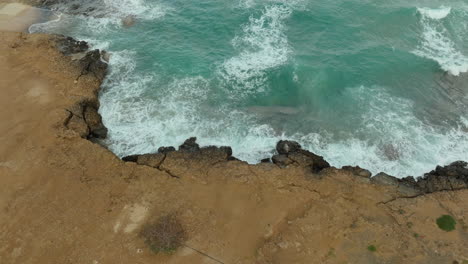 A-top-down-view-captures-the-dynamic-interaction-of-waves-crashing-against-a-rocky-shoreline,-surrounded-by-sandy-terrain-and-the-vivid-blue-of-the-sea