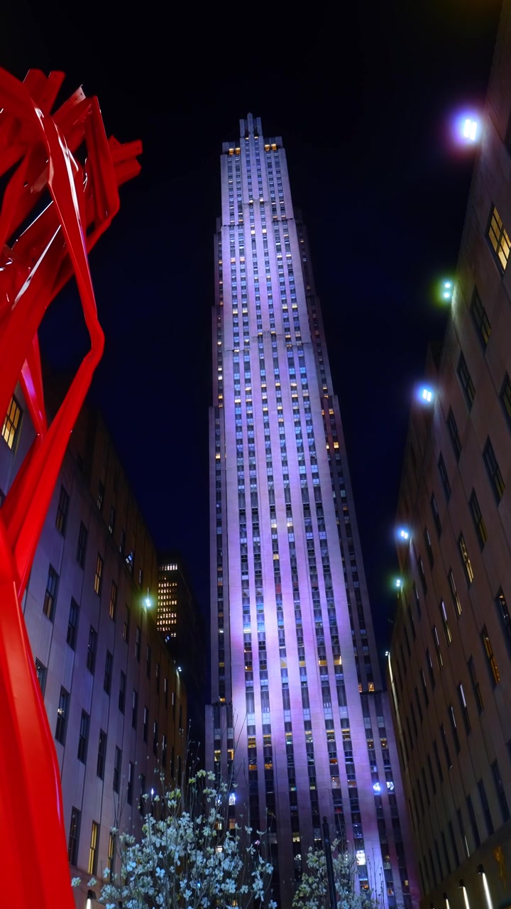 Grattacielo illuminato di notte, Rockefeller Center, New York City. Vista verticale dal basso di un'alta torre che brilla di viola con una scultura rossa e alberi in fiore bianchi in primo piano.