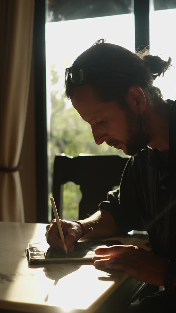 A man is drawing at a table near a window