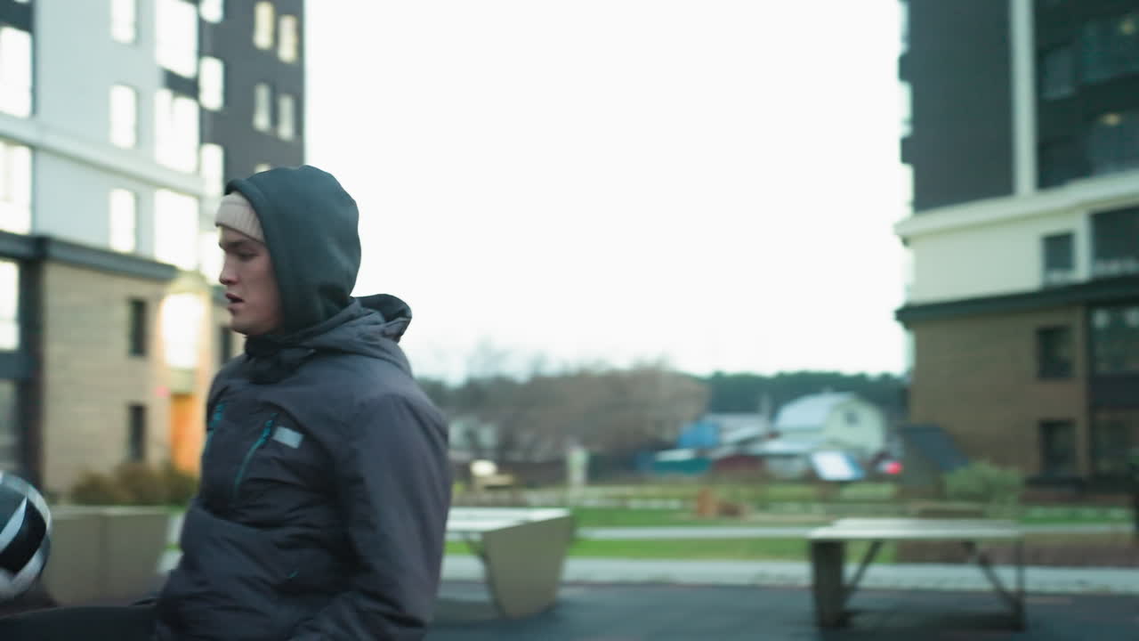 Sports man juggling ball during training in urban sport arena, with benches, buildings, fitness equipment, and blurred people walking in the background