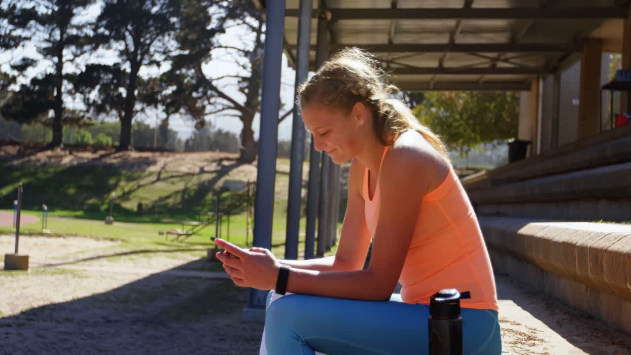 vista lateral de una atleta caucásica usando un teléfono móvil en un recinto deportivo 4k