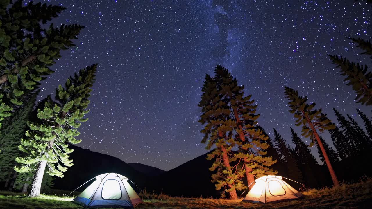 A wide-angle video shot of illuminated tents under a starry night sky, framed by tall pine trees