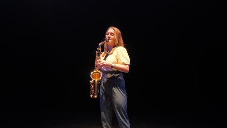 young woman musician playing solo on saxophone under lights on dark stage