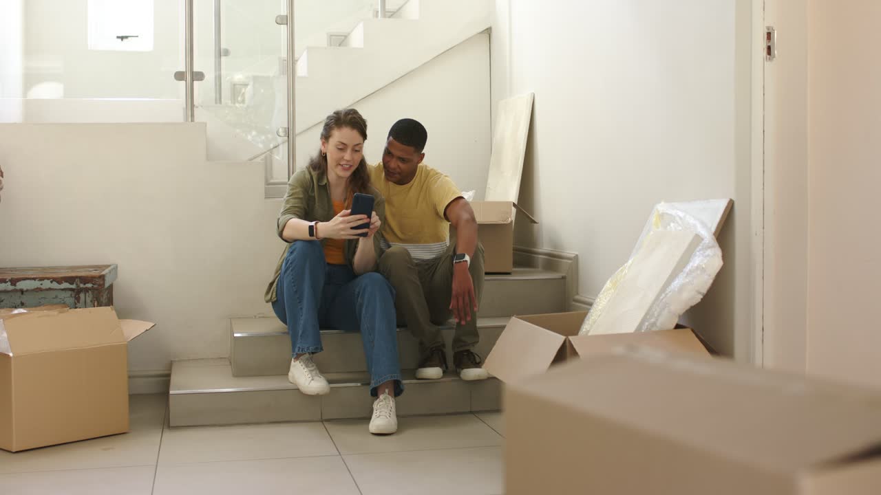 Couple resting among moving boxes on stairs, man carrying lamp descending, joining woman at phone