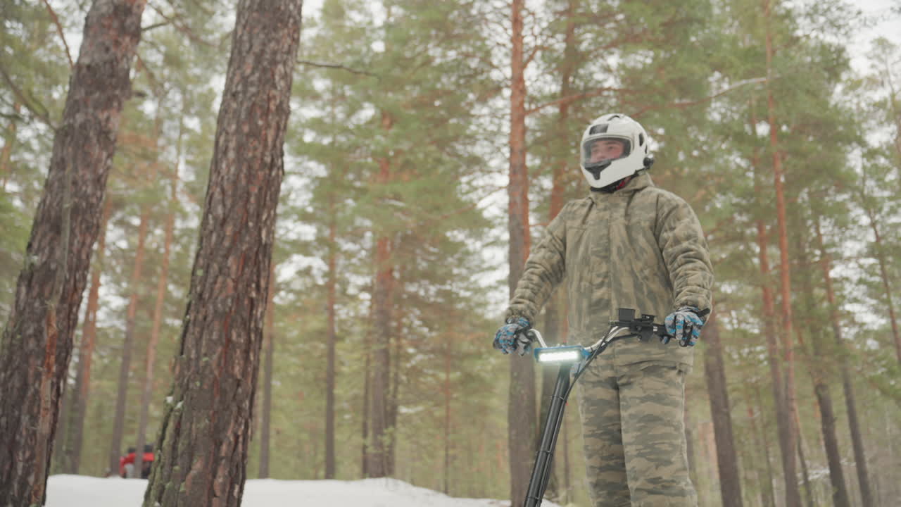 Pilota di quad con casco nei boschi innevati, in piedi al manubrio con faro luminoso, indossa giacca isolante e occhiali, stringendo l'acceleratore e preparandosi a scivolare nella neve tra alti pini, polvere che si solleva, luce fredda e pallida.