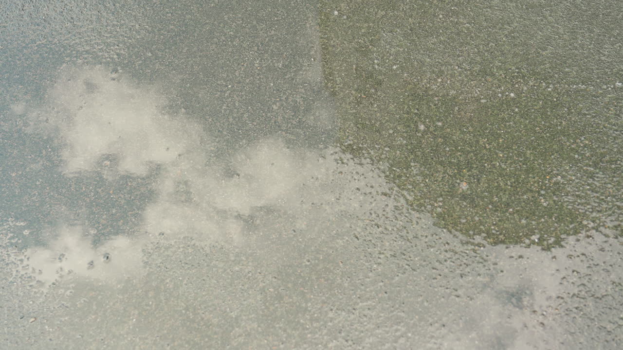 Rainy puddle reflecting cloudy sky, turf, slow ripples from steady drizzle, scattered raindrops creating micro splashes, surface sheen capturing muted stadium silhouette, wet grass edging, moody
