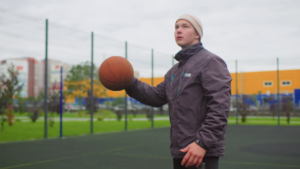 young white man on outdoor court dribbling and shooting basketball in beanie and padded jacket during overcast autumn day showing focused warmup, precise aim, jump shot, and reflective gaze