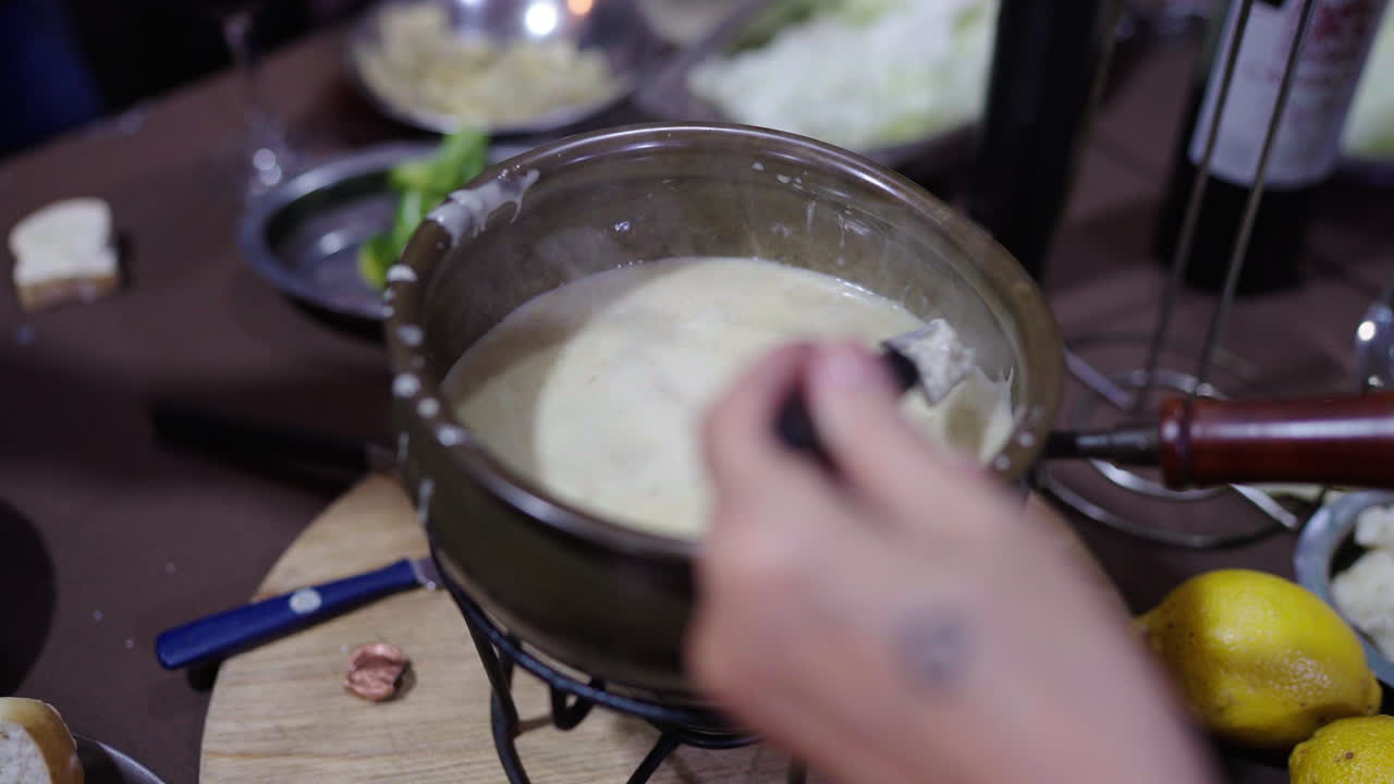 Creamy pot of Bagna Cauda sits at the center of a rustic table as one fork dips meat and another pasta, highlighting the Piedmontese tradition of communal dipping in garlic-anchovy sauce.