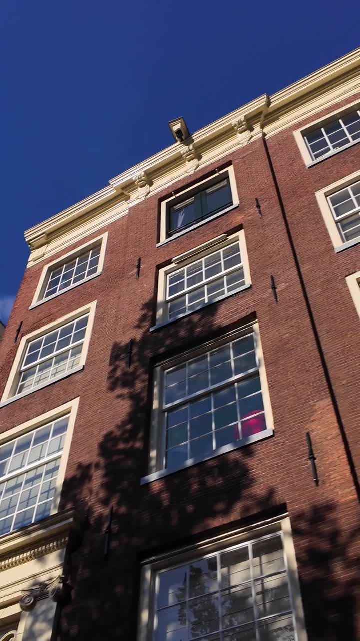 Historisk Amsterdam kanalhus facade med karakteristisk gavl og mange store vinduer under en klar blå himmel på en lys solskinsdag. Karakteristisk murstensarkitektur viser skygger fra