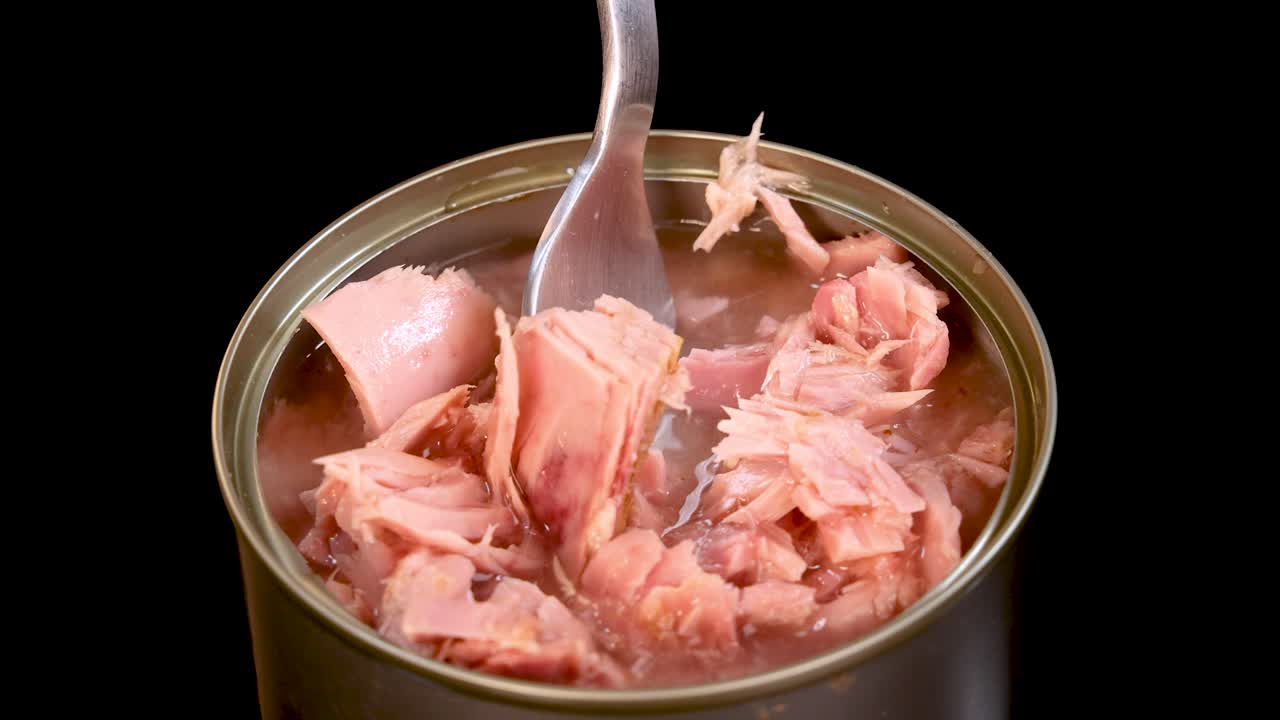 A fork lifts a chunk of tuna from an open can filled with mineral water, under bright studio lighting with a static, close-up camera angle