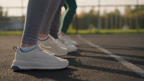 vista lateral de las piernas de personas con ropa deportiva y zapatos de lona saltando y moviéndose lateralmente con la luz del sol reflejándose en ellas, instalaciones deportivas visibles en el fondo lejano