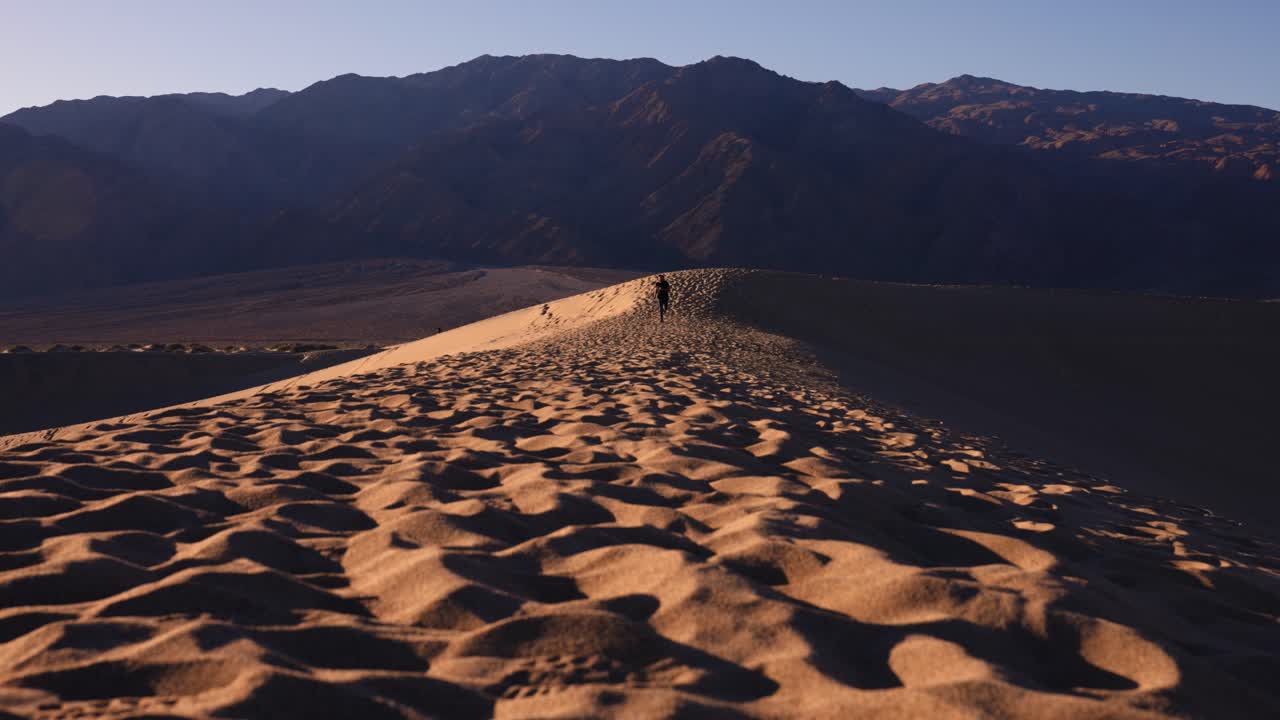 백그라운드에서 큰 산과 화창한 날에 캘리포니아 데스 밸리 국립 공원에서 모래 언덕 위에 카메라를 향해 slowmotion에서 실행 성인 남성