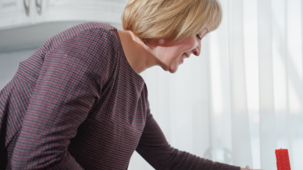 mujer de mediana edad coloca suavemente una vela roja en la mesa de la cocina en una cocina moderna brillante, armarios blancos, cortinas blancas transparentes y un ambiente hogareño acogedor