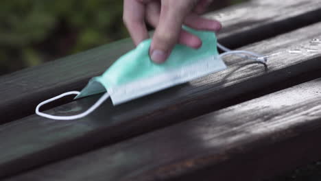 a simple green protective covid-19 face mask lying thrown out on a dark wooden bench after rain, a person picking it up carefully, blurry bushes in the background, static close up 4k shot