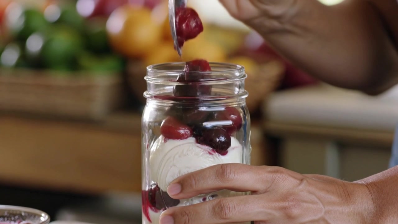 Mani che preparano un dessert, tenendo un barattolo di vetro sul bancone e usando un cucchiaio per stratificare crema e ciliegie.