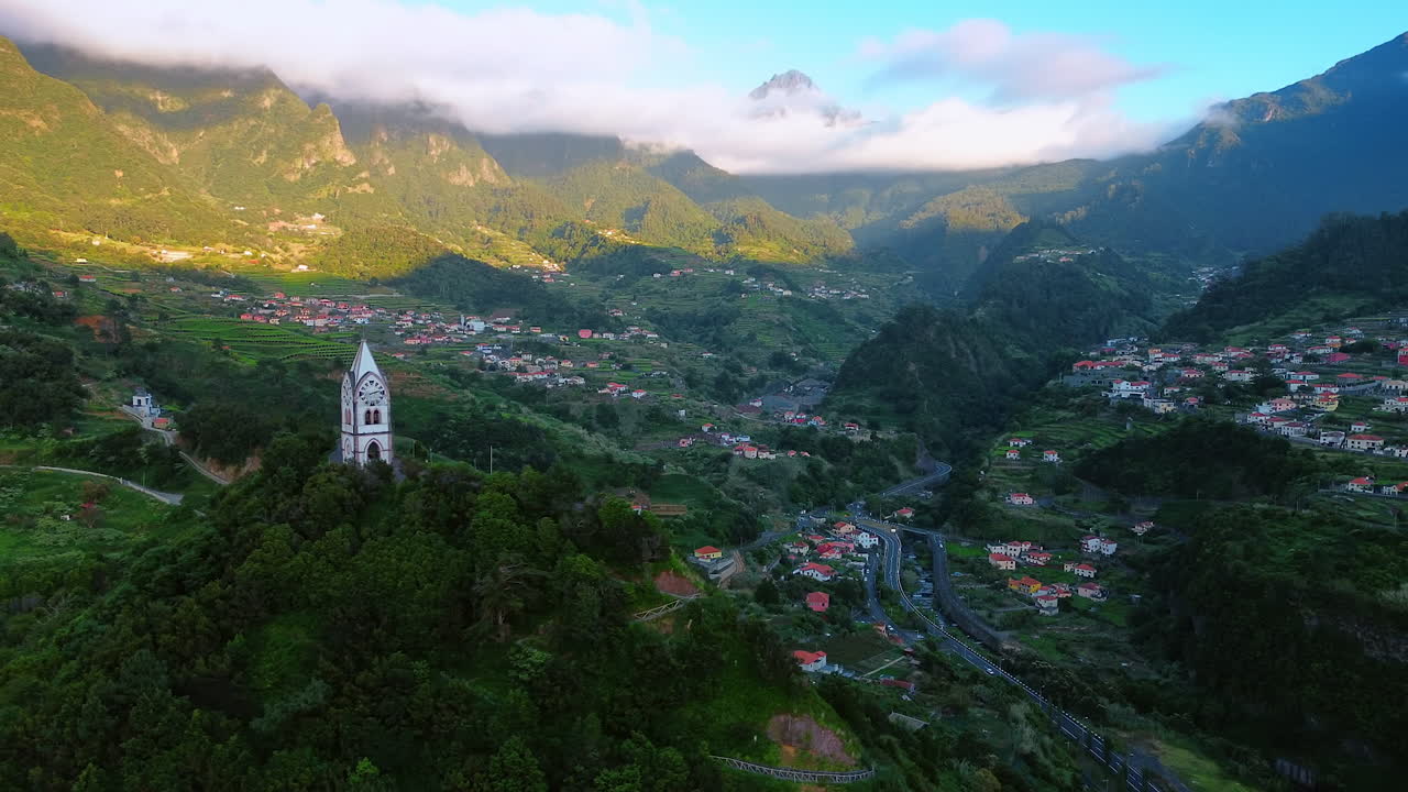 White tower clock located on the mount overgrown with lush greenery. Drone footage revealing the view on the picturesque town among the high mountains. White cloudscape covering the tops of the rocks.