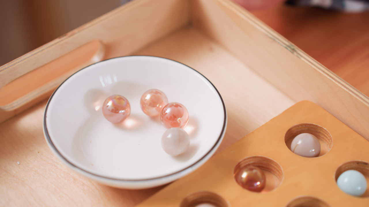 Close up toddler hand using tool to pick shiny balls from bowl and place them into wooden puzzle box, practicing coordination and focus during playful learning activity on smooth wooden surface