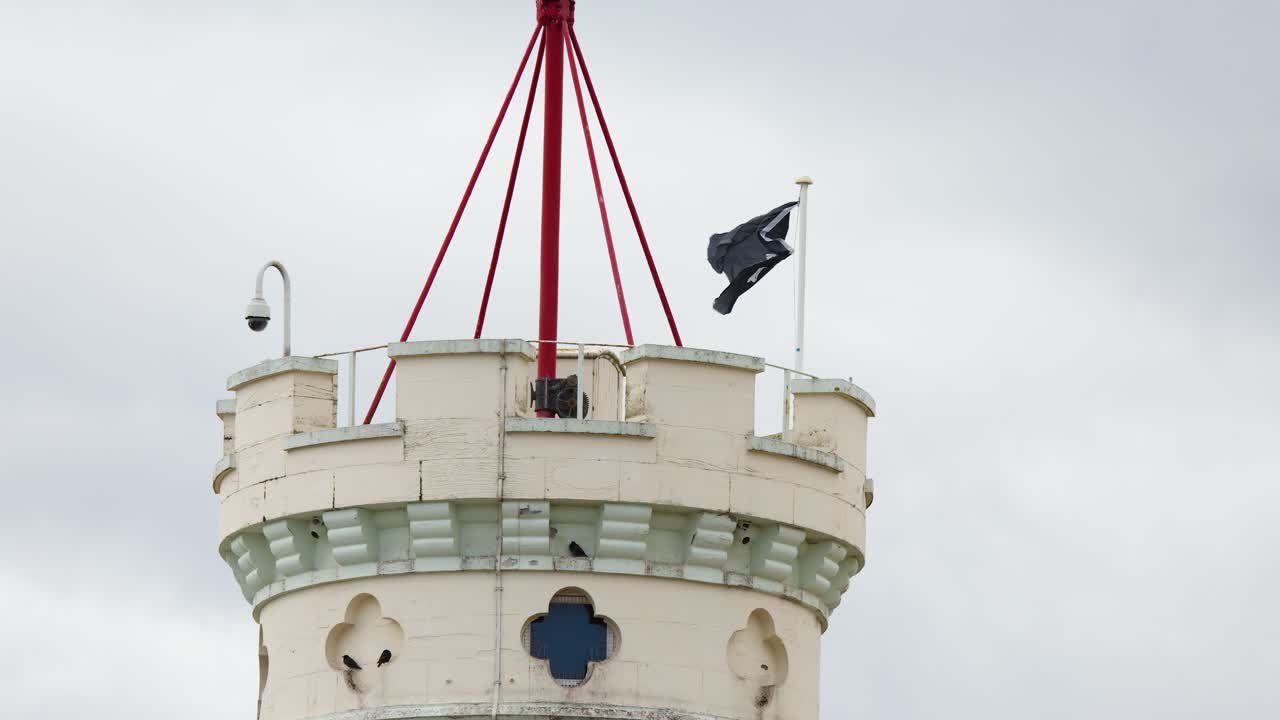 Black pirate flag waves atop stone tower under cloudy daylight, static camera, overcast ambiance