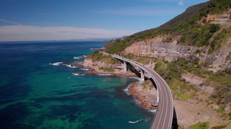Aerial view of cars driving on Sea Cliff Bridge, sunny day, grand pacific drive, New South Wales, Australia - dolly forward drone shot