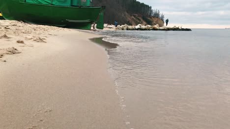 Paseo-Tranquilo-A-Lo-Largo-De-La-Orilla-Del-Mar,-Suaves-Olas-Golpean-La-Orilla,-En-La-Distancia-Se-Puede-Ver-Un-Barco-De-Pesca-Verde-Y-Piedras,-En-El-Fondo-Un-Acantilado-En-El-Fondo,-Cámara-Lenta-En-4k