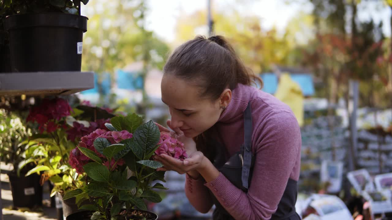 joven hermosa y sonriente jardinera acariciando y olfateando flores de hortensia en el invernadero mientras trabaja, vista en primer plano