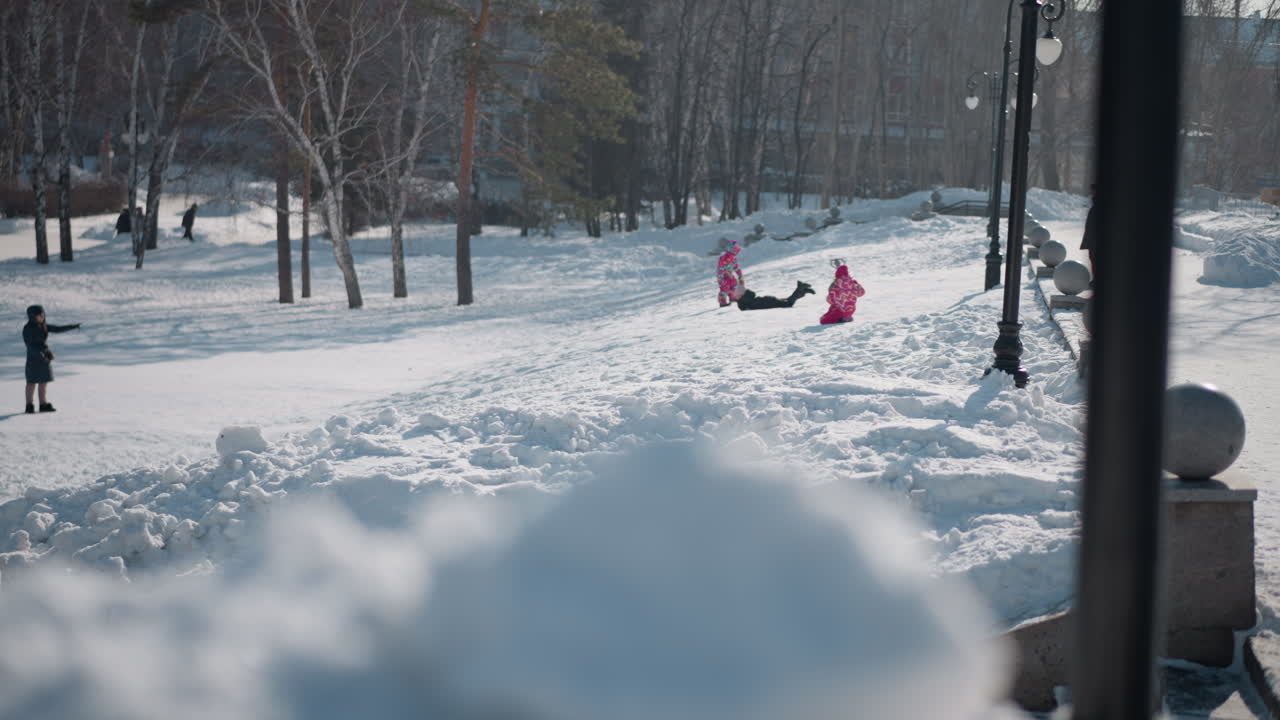 Kid sliding down high snowy walkway while adults watch from park path, lamp posts and granite spheres lining route, birch and pine trees around, bright winter daylight, blur foreground snow mounds
