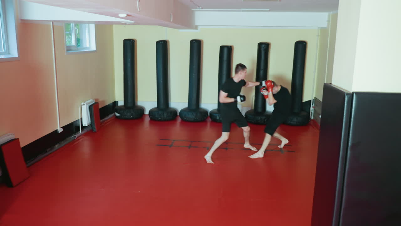 레슬링 선수들이 격투기 체육관 안 붉은색 매트 위에서 맨발로 글러브와 검은색 운동복을 입고 강렬하게 전투 기술을 연마하고 있어요. 주변에는 샌드백과 부드러운 벽 패딩이 있네요.