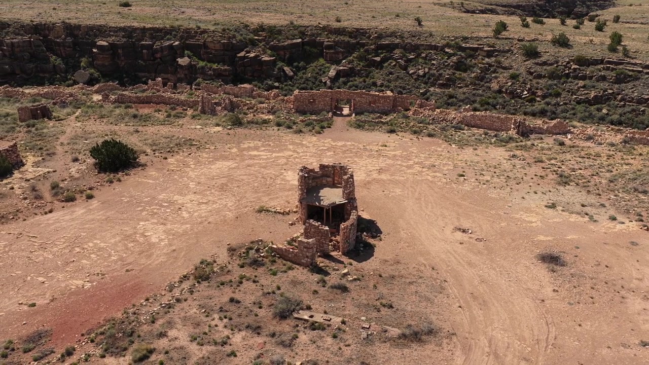 A cinematic drone orbit captures the abandoned stone ruins and ghost town structures of Two Guns, Arizona. The rotating camera moves around the weathered walls and roofless buildings perched on the ed