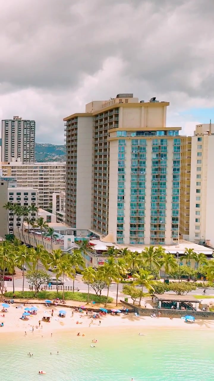 Vista aérea de una torre de hotel beige en Waikiki con paneles de cristal verde azulado, palmeras, sombrillas de playa, turistas y océano turquesa. Video vertical.