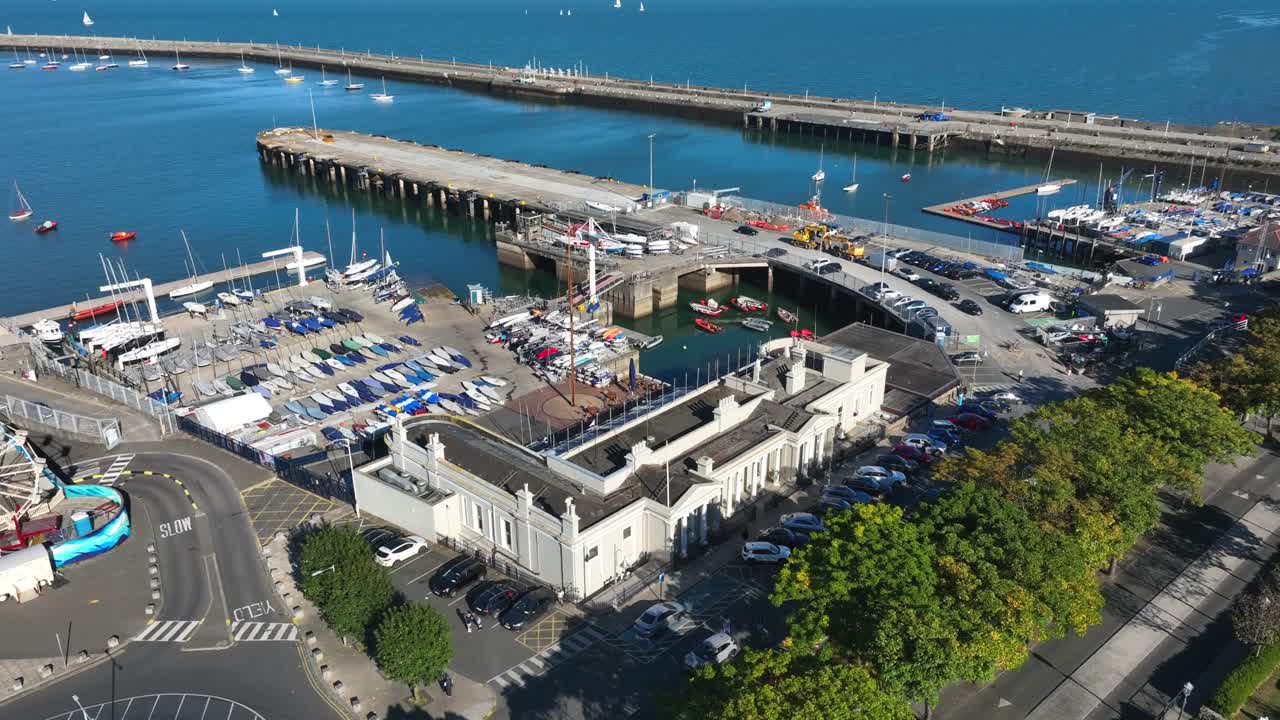 Royal St. George Yacht Club, Dún Laoghaire, County Dublin, Ireland, September 2024. Drone orbits counter clockwise while ascending above tree lined coastal road and plaza with small fun fair rides.