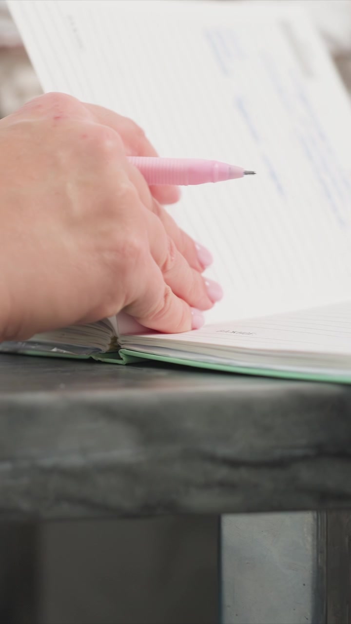 Primo piano della mano di una donna che tiene e scrive con una penna rosa in un quaderno aperto su una superficie di marmo. La scena mostra un'illuminazione soffusa e costante e una calma attenzione al movimento preciso delle dita sulle righe.