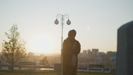 close-up shot of a sad girl wearing a coat and scarf, her hair partially covering her face, as she stands in front of a stone monument in a park during sunset