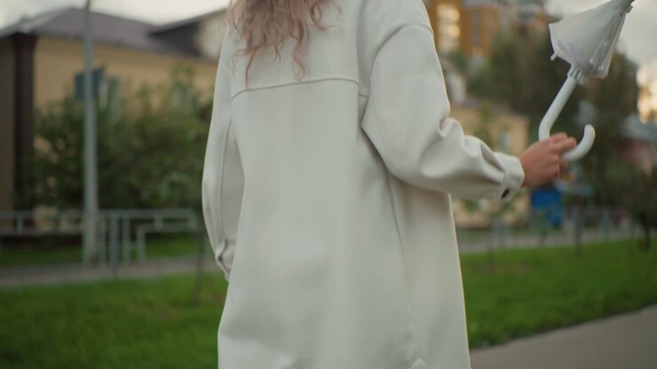 Rear view of woman in long white coat walking to work while twirling closed white umbrella, carrying handbag, soft blurred view of residential building, iron fence, and greenery in urban background