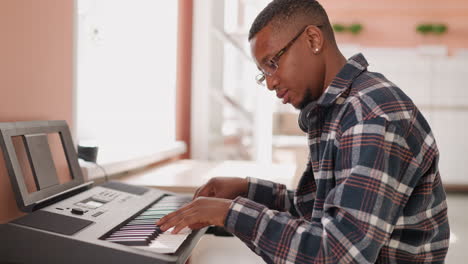 music creator writes composition on synthesizer. black man develops musical ideas using electric device at home. african american guy honing musical talents