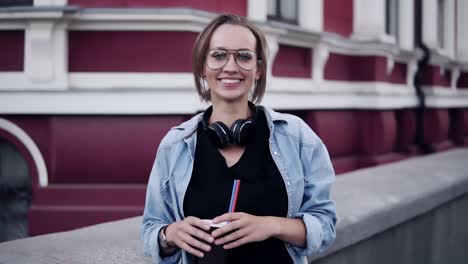 Gorgeous-View-Of-A-Young-Beautiful-Girl-In-Glasses-Standing-With-A-Red,-Classic-Building-On-The-Background-And-Smiling-To-Camera