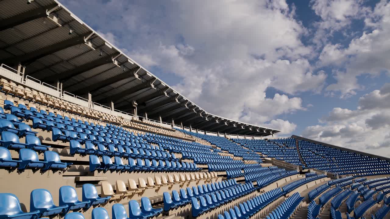 Rows of empty blue and beige stadium seats create a wave like pattern beneath a cloudy sky, evoking feelings of anticipation and the vastness of an open air venue