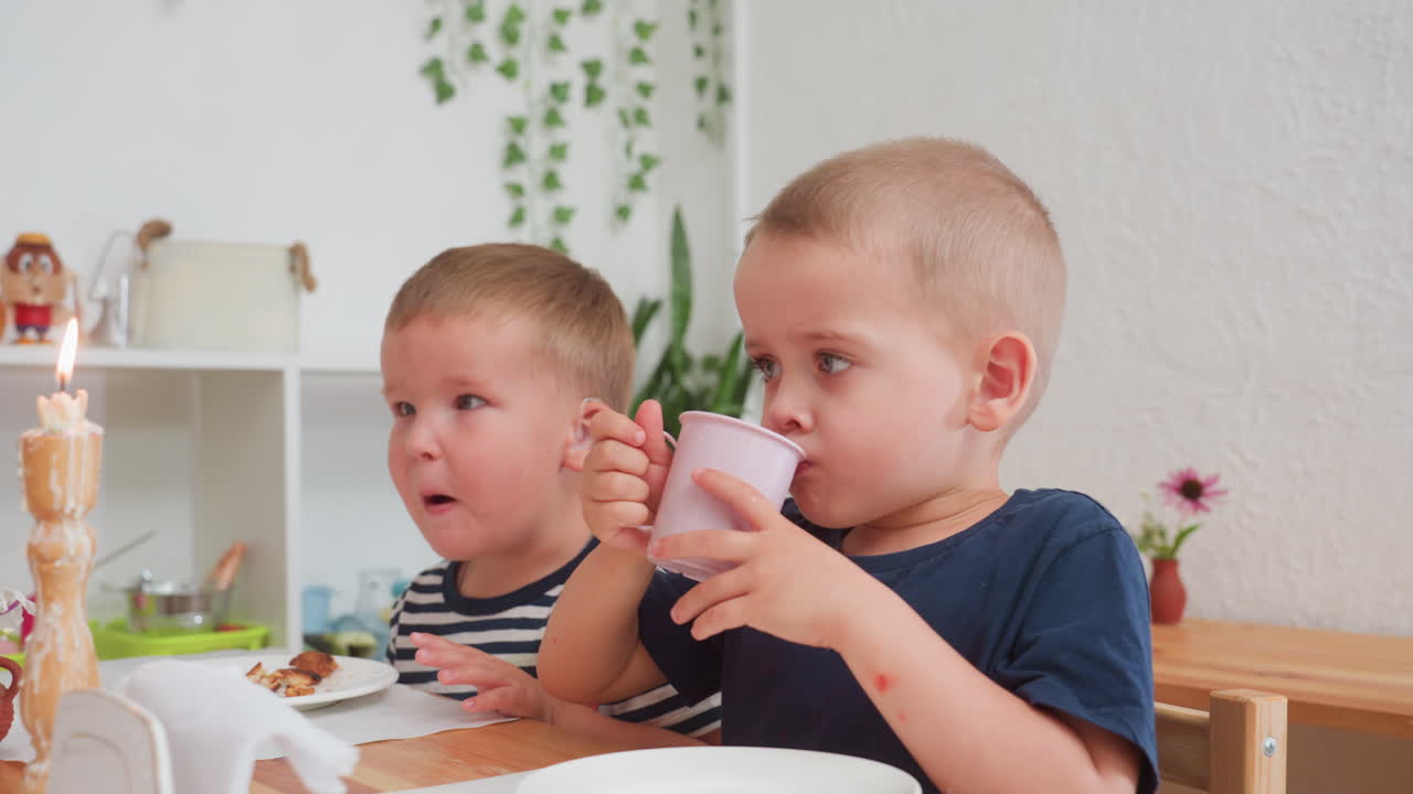 Two toddlers sitting at wooden table during kindergarten meal, one drinking from pink cup while other looks attentively surrounded by candle, plates, and plants under soft daylight
