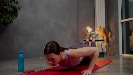 confident athletic brunette girl in a purple top and gray sweatpants on a red sports mat does sports runs in place and does push-ups in a modern apartment near a large indoor plant