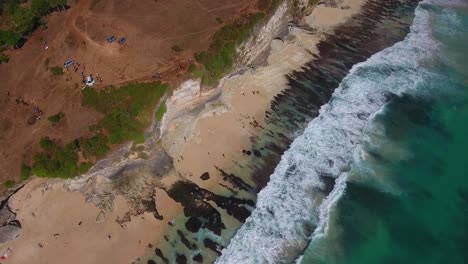 looking-down-view-of-waves-rolling-in-to-rocky-beach-with-big-cliffs,-uluwatu,-bali,-indonesia