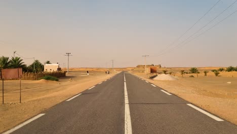 shot of an empty highway in the sahara desert near the town of timimoun, algeria. people walking in the background, with palm trees and houses along the roadside on a hot sunny day