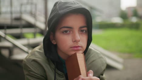 adolescent in hooded jacket sits on bench outdoors resting chin on hands holding brown book, staring calmly with composed expression against blurred background of urban greenery and stairs