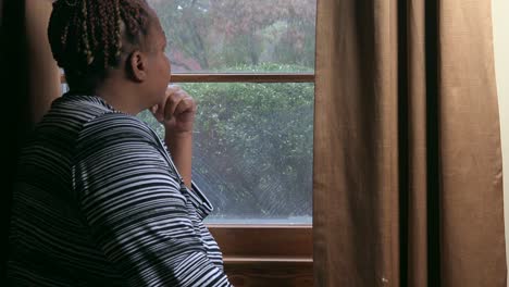 Mujer-Negra-Sentada-Junto-A-La-Ventana-De-La-Sala-De-Estar-Mirando-Mientras-Llueve-Esperando-Días-Mejores-Por-Delante